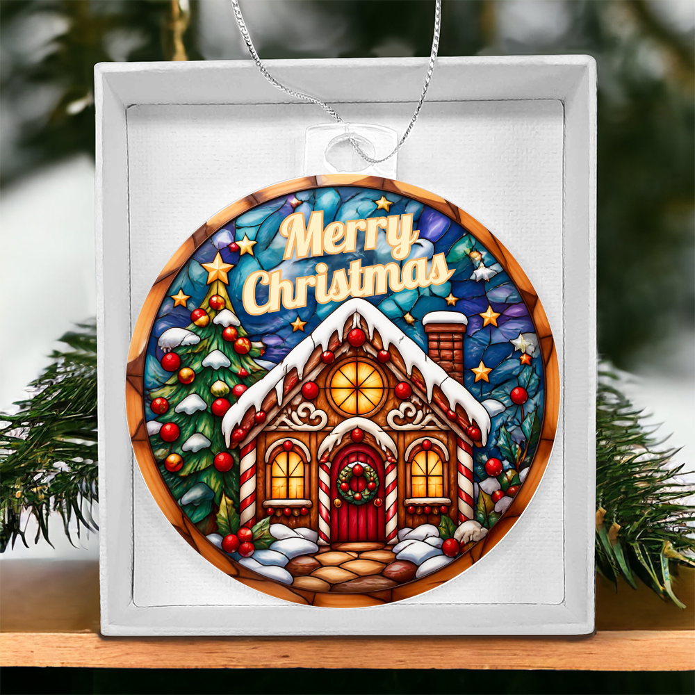 Round acrylic Christmas ornament in a stained-glass style featuring a illuminated gingerbread house surrounded by snow-covered pine trees with "Merry Christmas" written at the top, displayed inside a white gift box on a wooden surface with pine branches in the background
