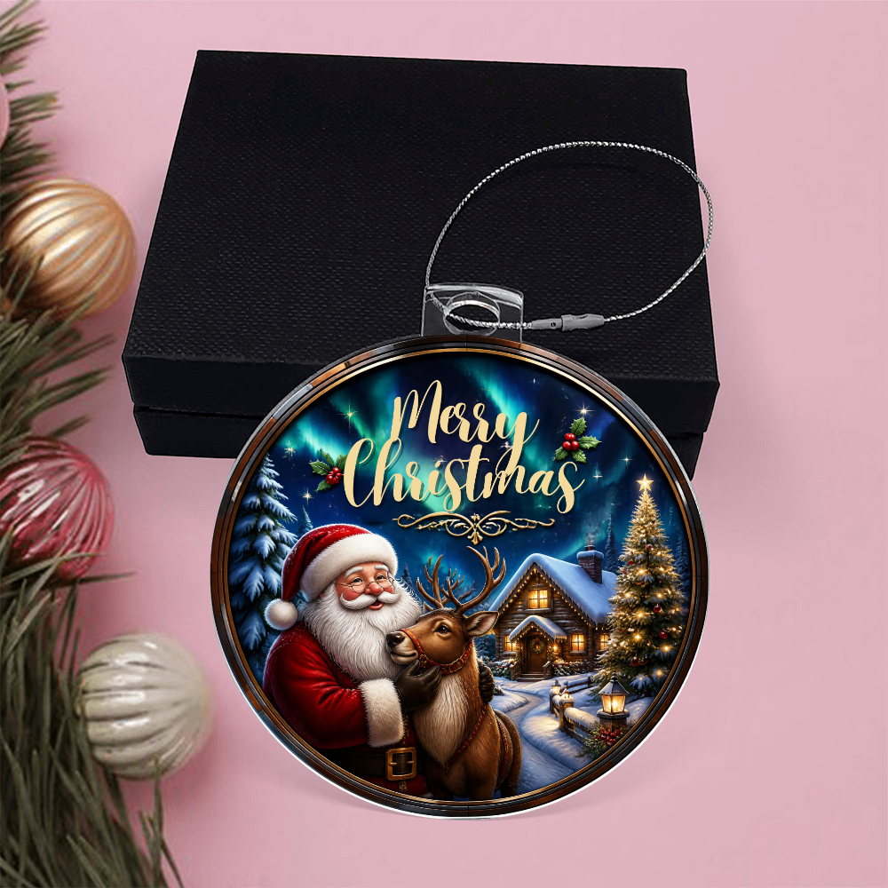 Round acrylic Christmas ornament in a stained-glass style featuring Santa Claus, a reindeer, and an illuminated winter cabin on a pink surface next to a black gift box.