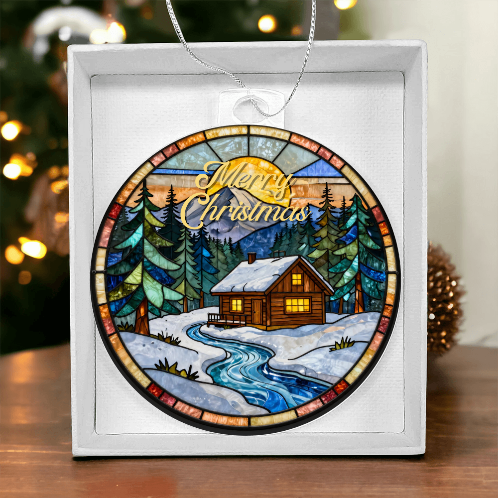 Round acrylic Christmas ornament in a stained-glass style featuring a cozy winter log cabin and pine trees, displayed inside a white gift box with blurred holiday lights in the background.