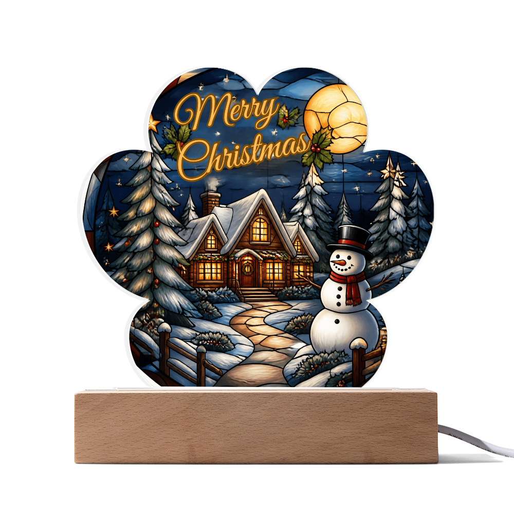 Paw-print shaped acrylic light-up ornament featuring a glowing winter cabin, snowman, and pine trees with "Merry Christmas" text, set on a wooden LED base.