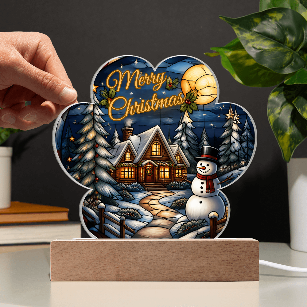 Hand placing a paw-print shaped acrylic light-up ornament onto a light wooden LED base, featuring a glowing winter cabin, snowman, and "Merry Christmas" text.