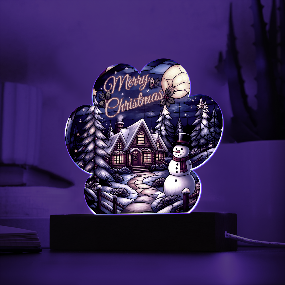 Paw-print shaped acrylic light-up ornament featuring a glowing winter cabin, snowman, and pine trees with "Merry Christmas" text, set on a dark wooden LED base.