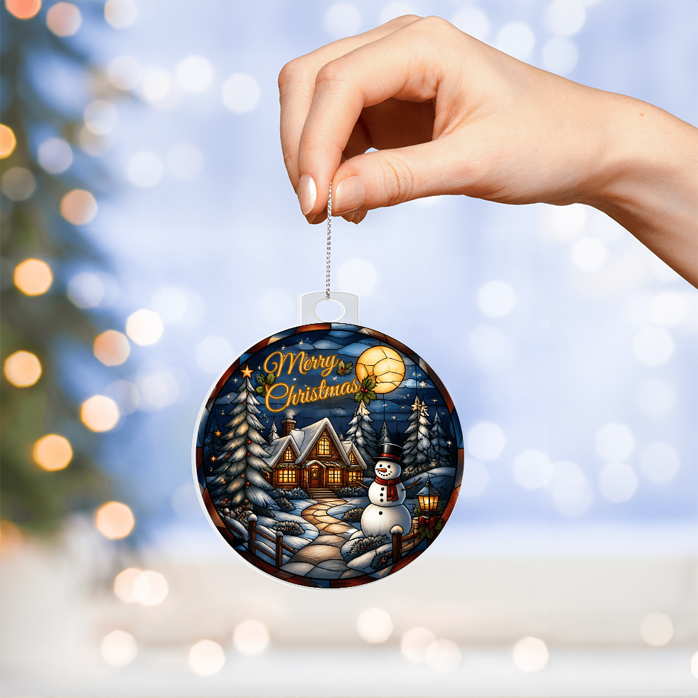 Hand holding a round acrylic Christmas bulb ornament featuring a glowing winter cabin, snowman, and pine trees under a full moon against a bokeh background.
