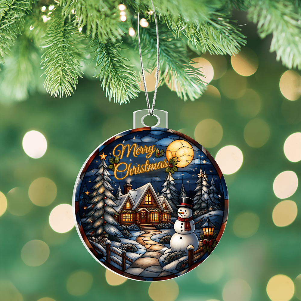 Round acrylic Christmas bulb ornament featuring a glowing winter cabin, snowman, and pine trees hanging on a Christmas tree.