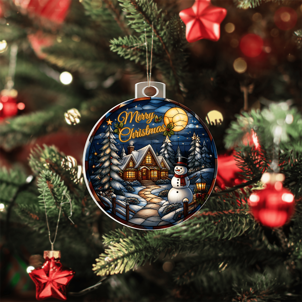 Round acrylic Christmas bulb ornament featuring a glowing winter cabin, snowman, and pine trees hanging on a Christmas tree.