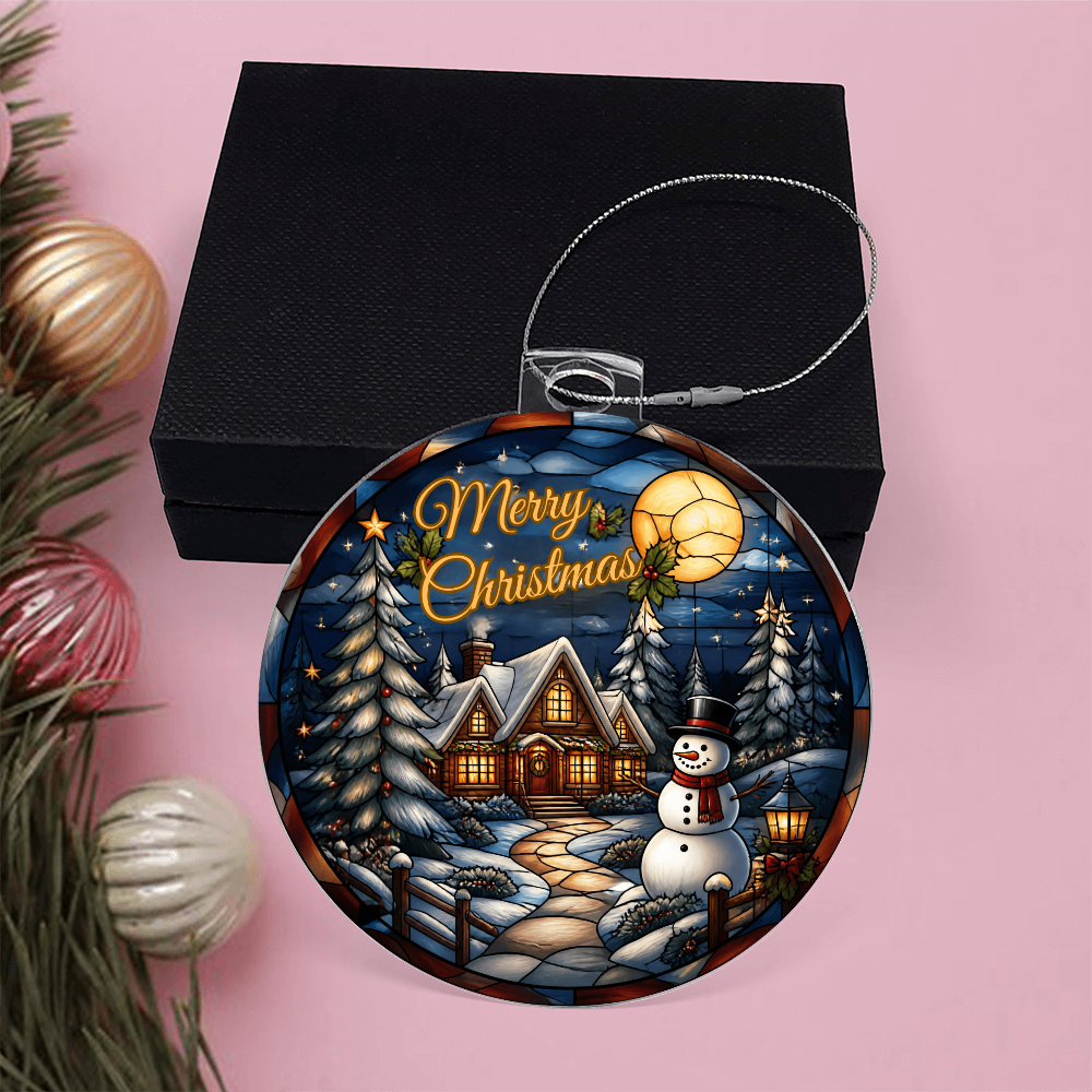 Round acrylic Christmas bulb ornament featuring a glowing winter cabin, snowman, and pine trees under a full moon, displayed next to a black gift box.