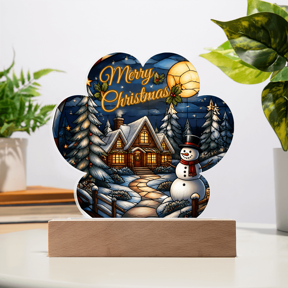 Paw-print shaped acrylic light-up ornament featuring a glowing winter cabin, snowman, and pine trees with "Merry Christmas" text, set on a dark wooden LED base.